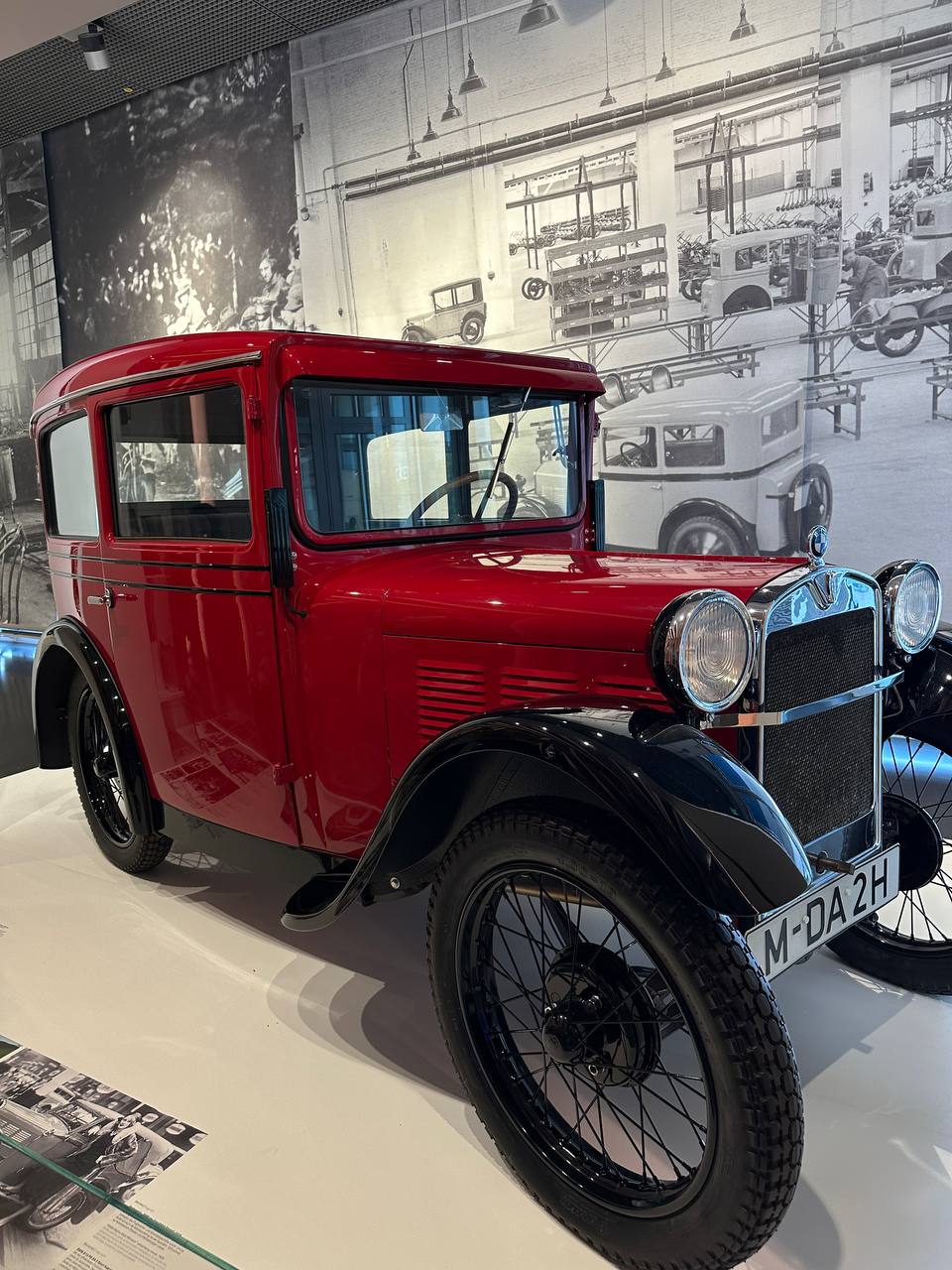 An early red BMW microcar displayed with historic factory imagery in the background.